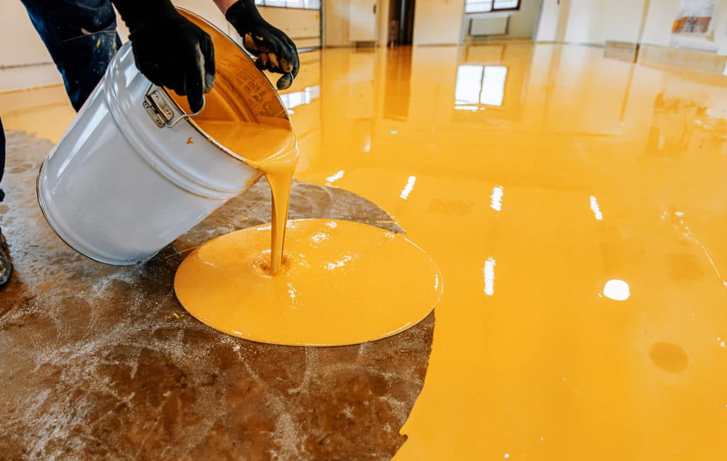 Person pouring epoxy floor coating in building.